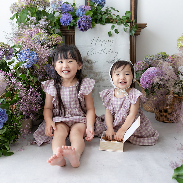 紫陽花で彩られたスタジオで幼い姉妹が仲良く横並びに座ってこちらに笑顔を向けている